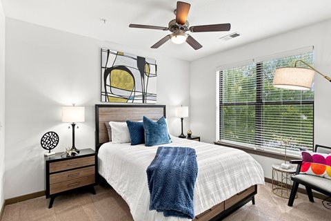 A bedroom with a bed, two lamps, a ceiling fan, and a painting on the wall.