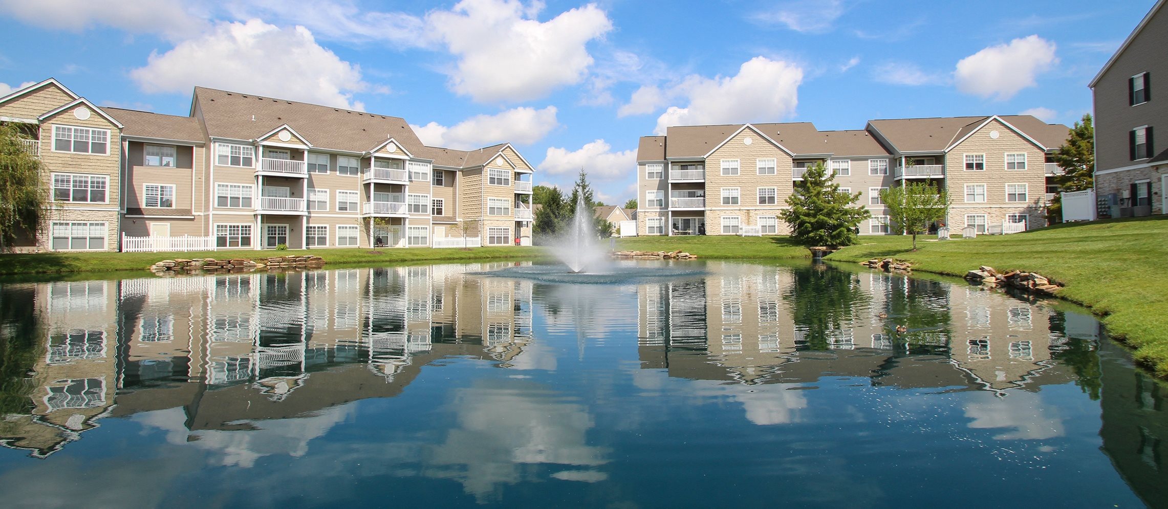 This is a picture of a fountain in a pond with building exteriors at Nantucket Apartments, in Loveland, OH.