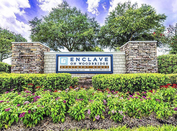 Stone pillar sign with a blue framed insert reading Enclave on Woodbridge Apartment Homes. The sign is surrounded by neatly manicured shrubs and flowers, in front of mature trees.