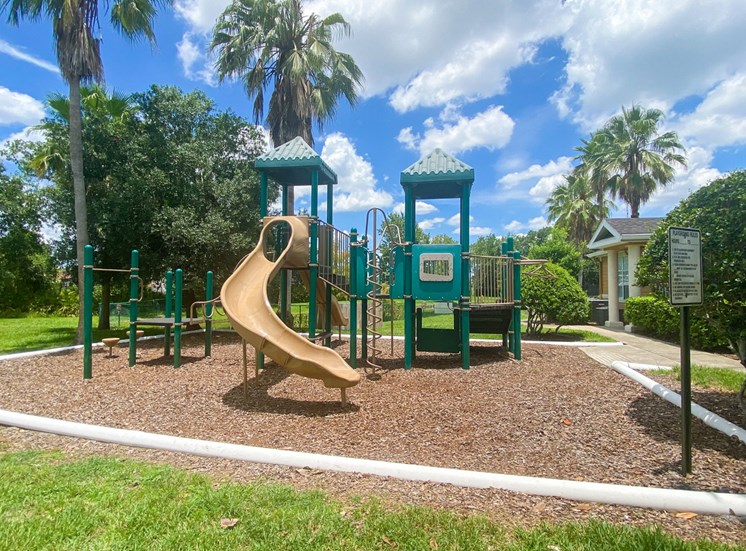 Green and yellow Playground set in a bed of mulch with buildings and palm tree, and bench in the background,