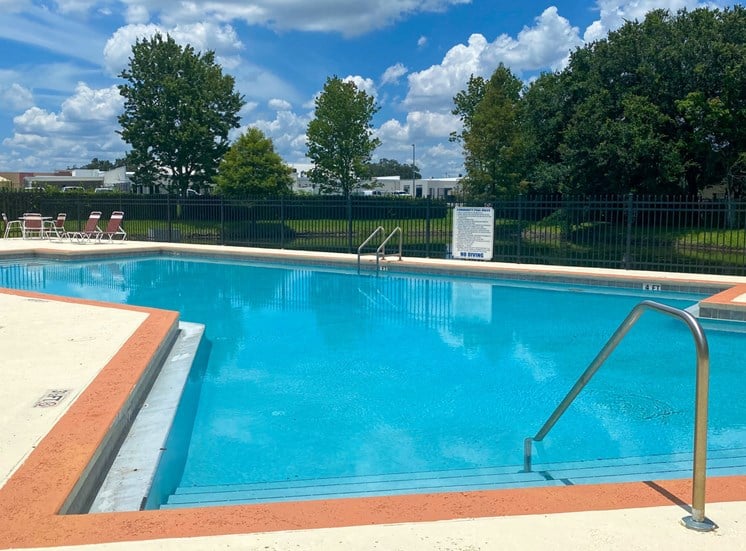 Community pool with sundeck, lounge chairs, surrounded by metal green fence with trees in the background