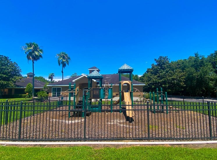 Green and yellow playground with jungle gym and slide in a bed of mulch surrounded by black metal fence with building exteriors and trees in the background