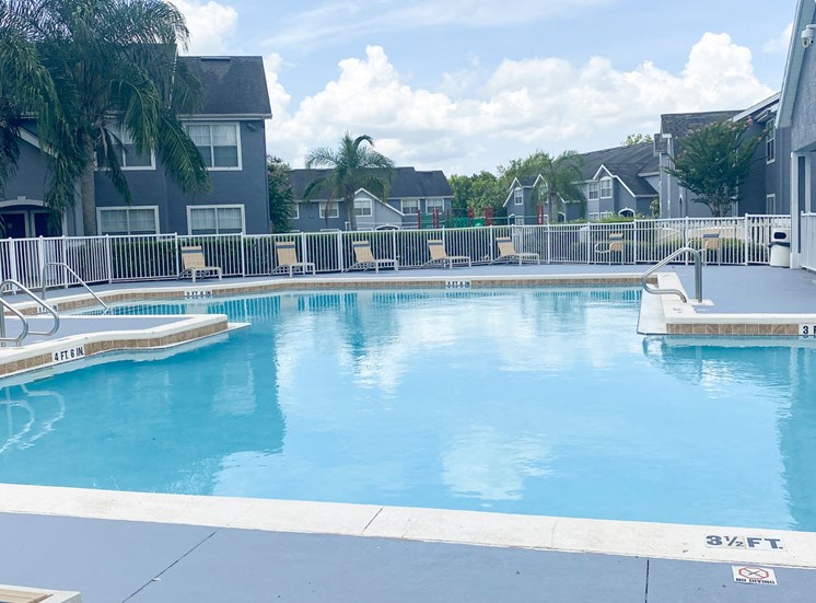 Community pool with sundeck, lounge chairs, with lake view surrounded by white metal fence with palm trees and building exteriors, palm trees, and trees in the background