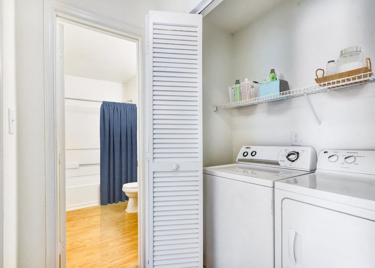A virtually staged laundry area with accordion doors, a full size washer and dryer, white walls and built-in shelving above the washer and dryer. It is staged with two baskets and cleaning supplies. The bathroom is located in the background.