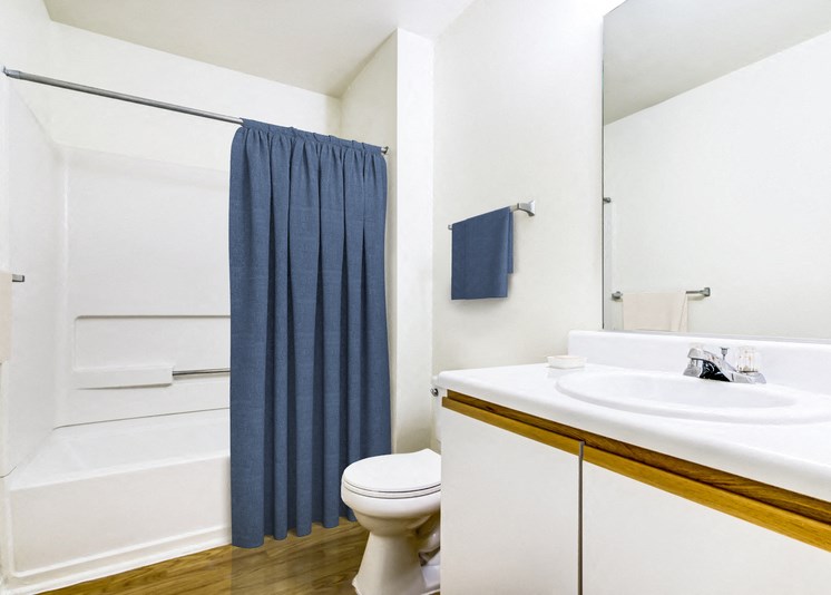 A virtually staged bathroom with hardwood style flooring, white painted walls, white countertops with single sink and white cabinets below, a single mounted mirror, one towel bar, one toilet and a shower/tub combo. It is staged with a navy blue shower curtain and matching towels