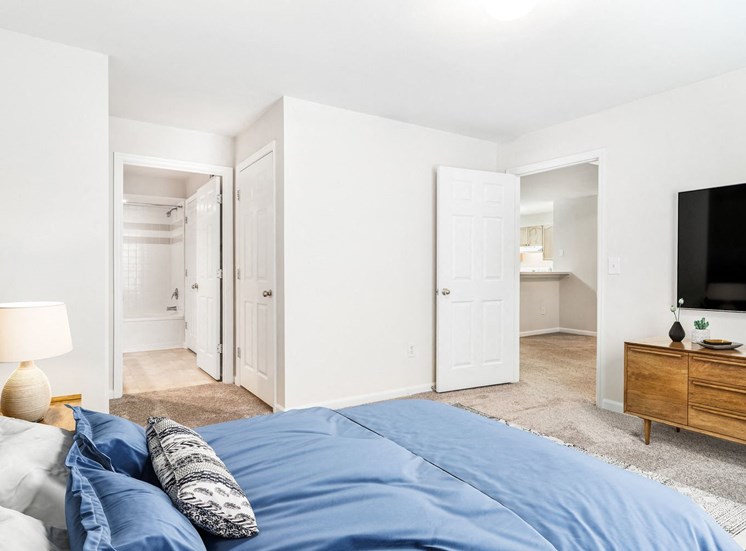 Vacant bedroom room with carpet. Nightstands on side of the bed, mounted tv above entertainment center with a view of the bathroom in the background.