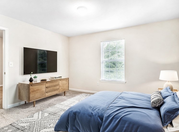 Vacant bedroom room with carpet. Nightstands on side of the bed, mounted tv above entertainment center with window.