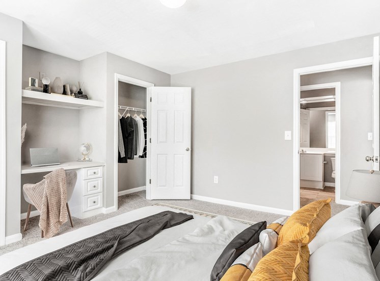 Staged bedroom room with carpet flooring and view of bathroom in the background. Clothes in open closet and décor above built in desk.