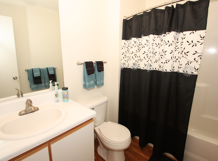 The furnished bathroom has hardwood style flooring, white with tan trim cabinetry under the sink, and a mounted mirror above the sink area. The wall behind the toilet has a towel rack with teal and black hand towels. The shower/tub combo has a black shower curtain with a teal and black floral pattern.