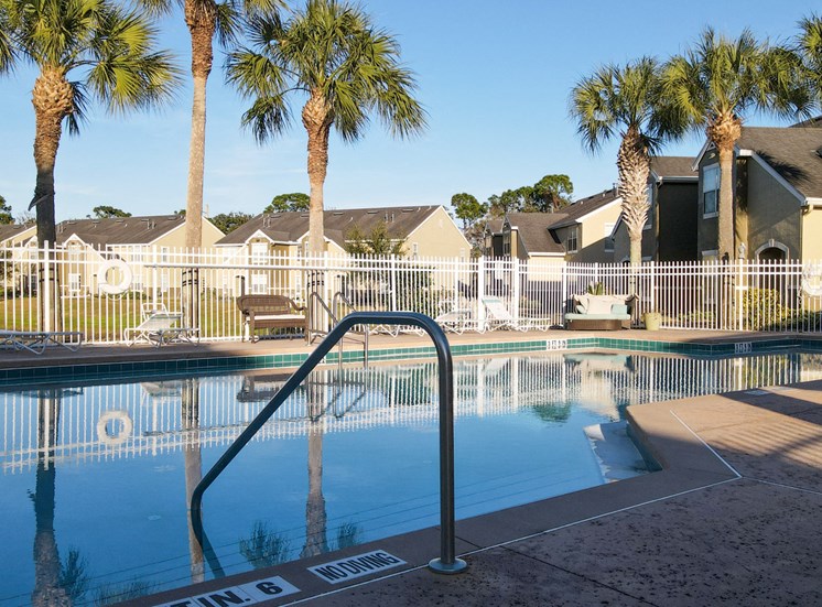 Community pool with sundeck, lounge chairs, with lake view surrounded by white metal fence with palm trees and building exteriors in the background