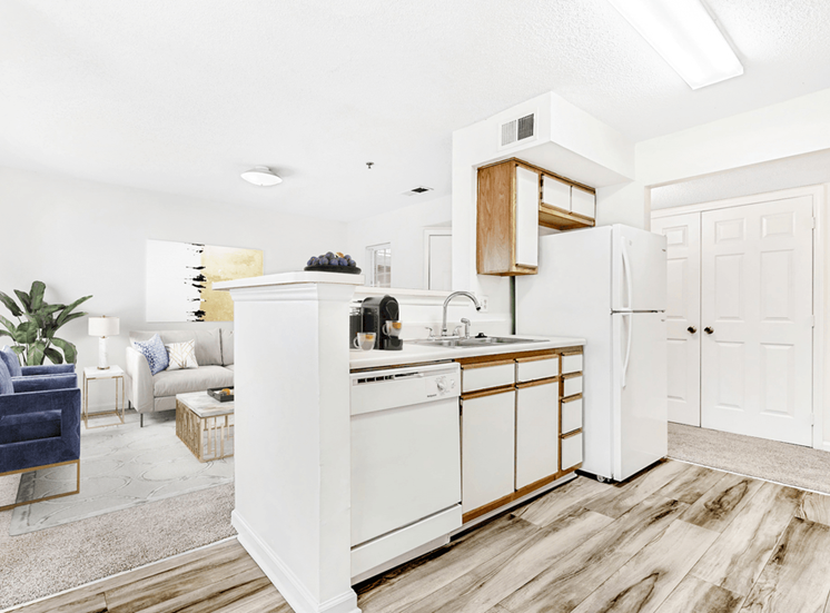 Virtually staged; Kitchen with wood floors, white appliances, and wood cabinets. Carpeted Living room in background with blue accent chairs