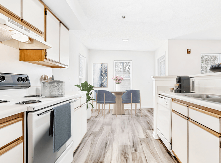 Virtually staged; Kitchen with wood floors, white appliances, and wood cabinets. Kitchen accessories and dining table in background with blue accent chairs