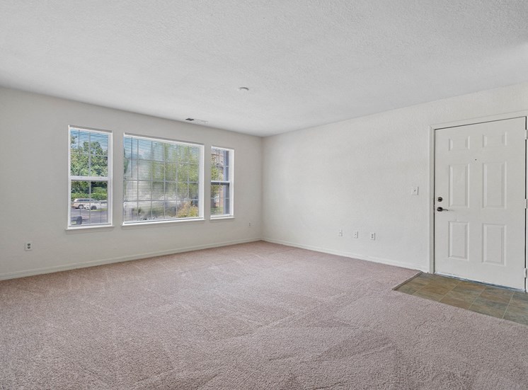 The living room features beige carpet, a linoleum tile foyer, and white walls. On the back left wall, there is a large picture window with two smaller windows on either side. The main entryway is located to the right in front of the linoleum tile foyer.