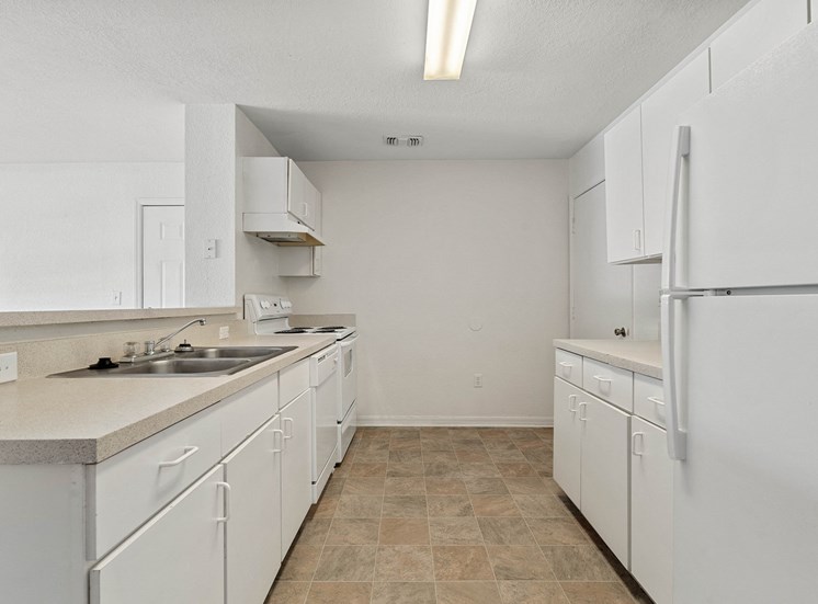 A vacant kitchen featuring white walls, linoleum tile flooring, white cabinets with white pulls, light colored formica counters, a double basin kitchen sink, and white appliances. Appliances include refrigerator, stove/oven combo, and dishwasher. There is a breakfast bar that opens to the living room.