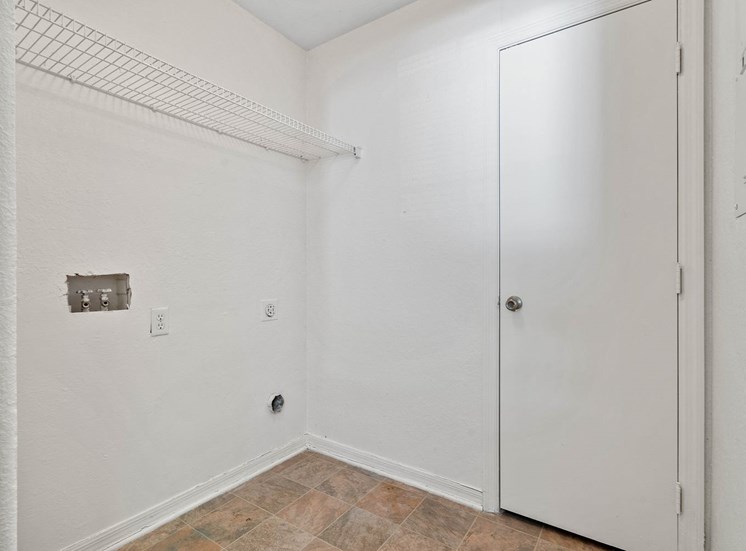 A vacant laundry room featuring washer and dryer connections below a white wire shelf that expands from corner to corner of the left wall. There are white walls and light beige linoleum flooring. A door that opens inward from the left is on the far wall with an electrical breaker panel to the right of the door.