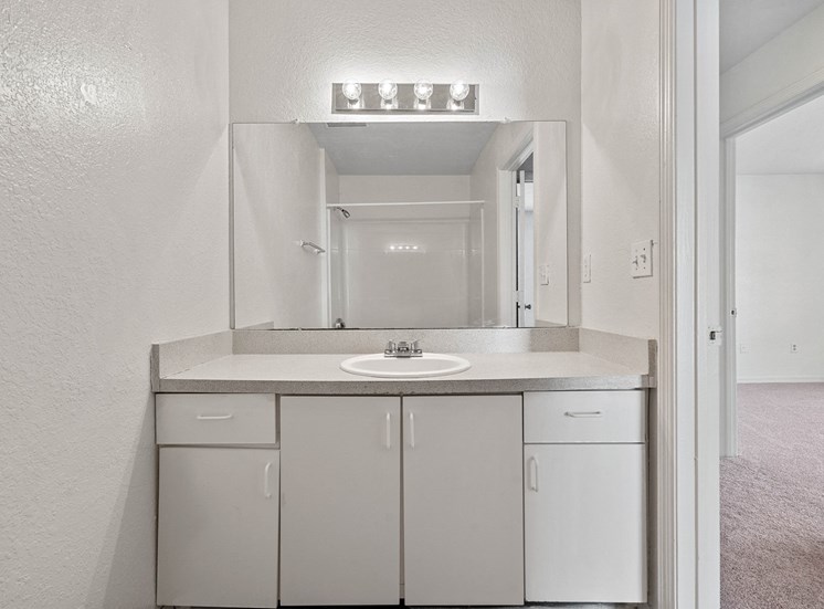 The second bathroom features white walls, a white vanity cabinet, a light-colored Formica countertop, and a white porcelain sink with chrome faucet. There is a large frameless mirror and chrome light fixture above the vanity.