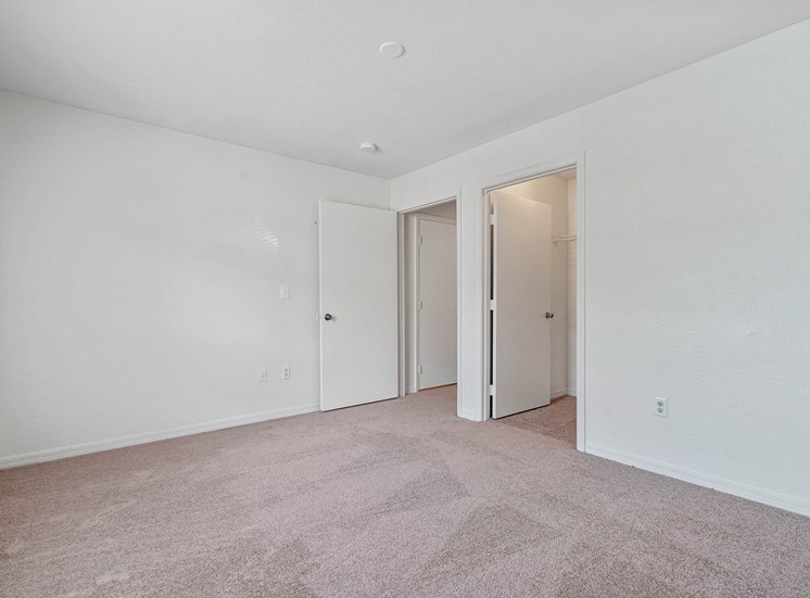 The vacant third bedroom features white walls and beige carpet. On the right wall is the doorway leading to the hallway. To the right of the entry door is the door to the walk-in closet. The walk-in closet has an overhead light and wired shelving.