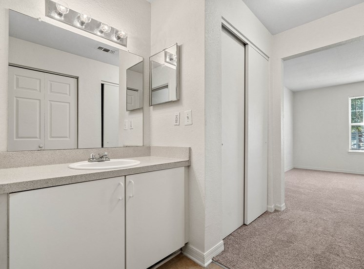 The bathroom connects with the bedroom. The walls are white and the carpet is beige. The vanity sits in an alcove and features a white cabinet with light colored countertop and white porcelain sink. Above the vanity there is a mirrored medicine cabinet on the right wall and a large frameless mirror above the sink with a chrome vanity light fixture.