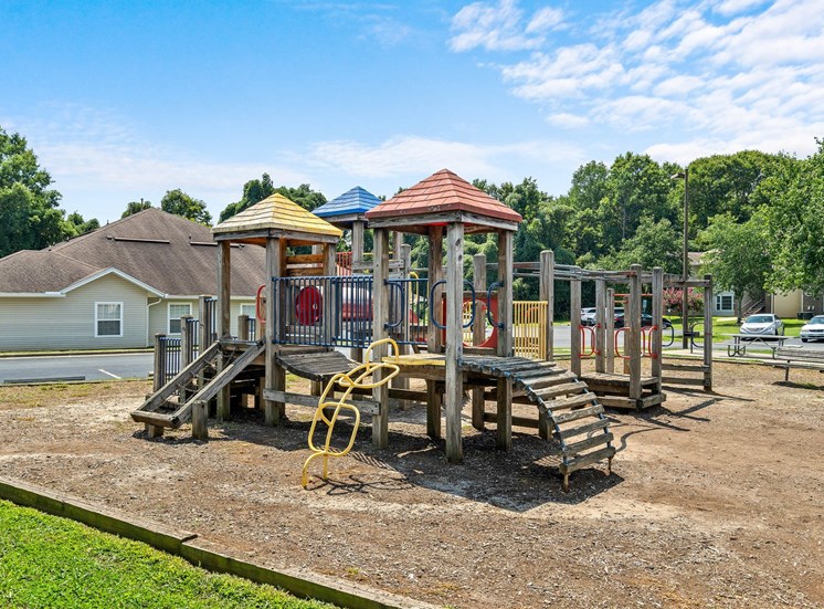 The community playground has a wooden base with red, yellow and blue accents. There is a red slide and several monkey bars. There is a bench in the background facing the playground equipment. The jungle gym and benches are surrounded by mulch with a wooden border. The exterior of the playground is surrounded by parking lots and apartment buildings.