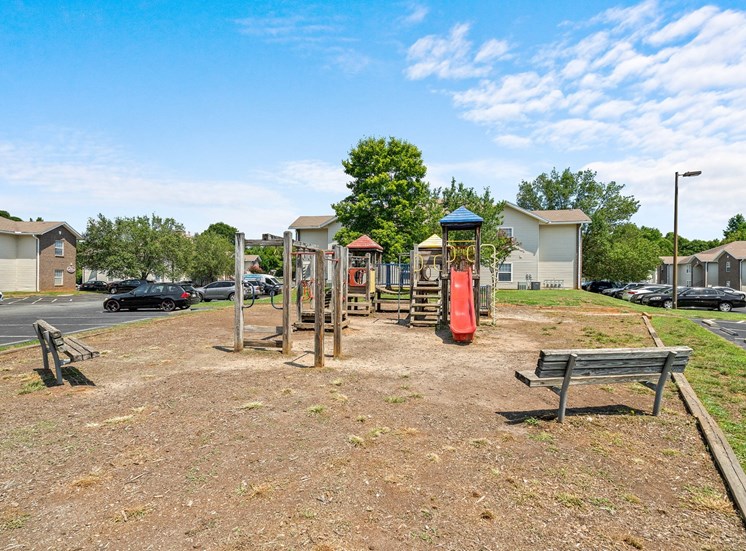 The community playground has a wooden base with red, yellow and blue accents. There is a red slide and several monkey cars. Two adjacent benches face the playground. The jungle gym and benches are surrounded by mulch with a wooden border. The exterior of the playground is surrounded by parking lots and apartment buildings.