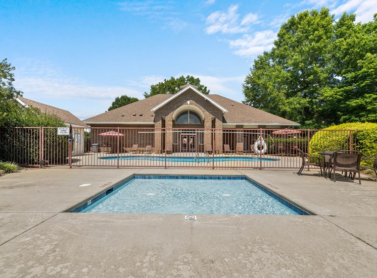 The wading pool is square and located in front of the swimming pool and clubhouse. The two pools are separated by a fence. Surrounding the wading pool is a concrete sun deck with a table and four chairs to the right. there are shrubs and trees around the pool decks and clubhouse.