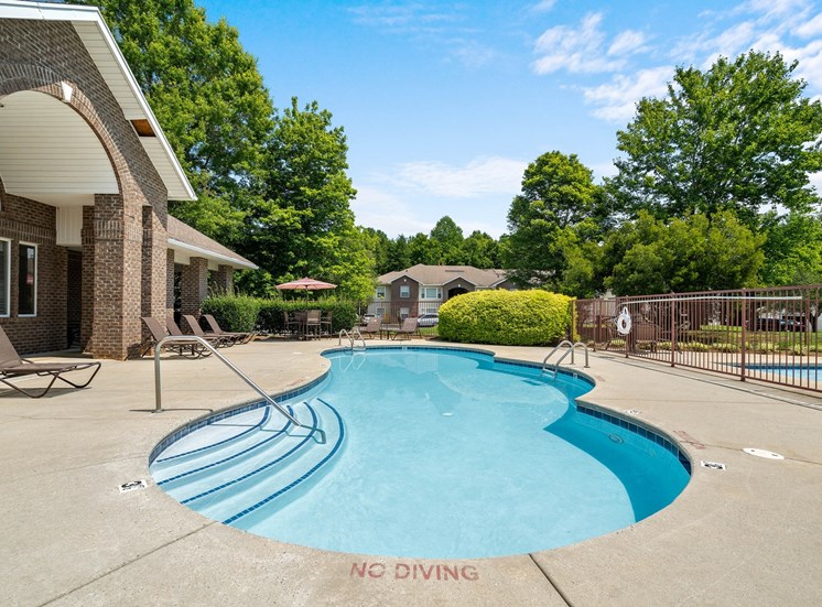 The oblong-shaped swimming pool is surrounded by a concrete sundeck. There are lounge chairs to the left in front of the Clubhouse. On the right is a fence separating the swimming pool from the wading pool. Outside the pool deck are trees, shrubs, and apartment buildings.