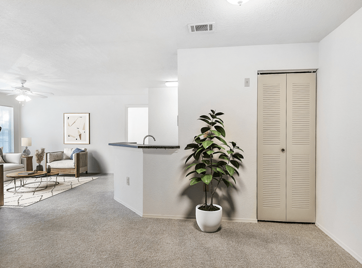 Virtually staged apartment entryway with carpet, indoor plants, livingroom with ceiling fan and light and breakfast bar in the background