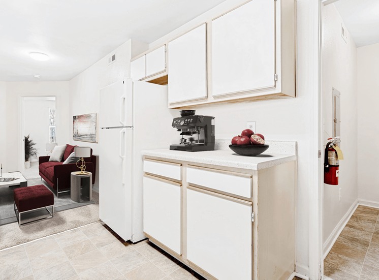 Virtually staged kitchen with tile floors, white appliances, and wood cabinets. Living room in background with maroon accent furniture.