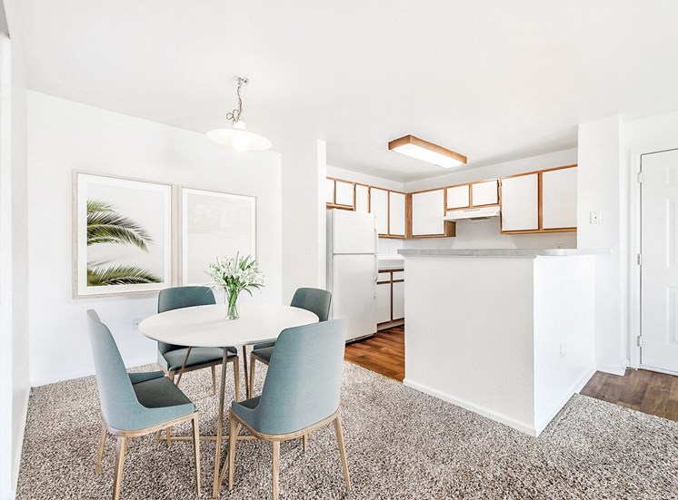 Model Apartment with Dinette Table in Front of Framed Wall Art Next to Open Kitchen with White Cabinets with Wood Accents and White Appliances