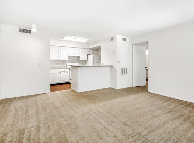 Vacant living room with wall-to-wall carpet. Kitchen in the background with white cabinets and appliances.