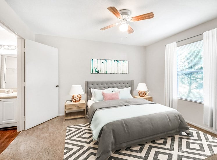 A staged bedroom with carpet, a ceiling fan, window, and an en suite bathroom. Full bed with tufted headboard on top of a rug and two night stands, and art above the bed.