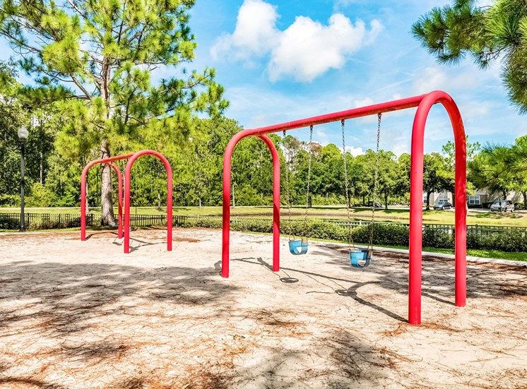 Two red swing sets in the playground area surrounded by an open grass area and trees.