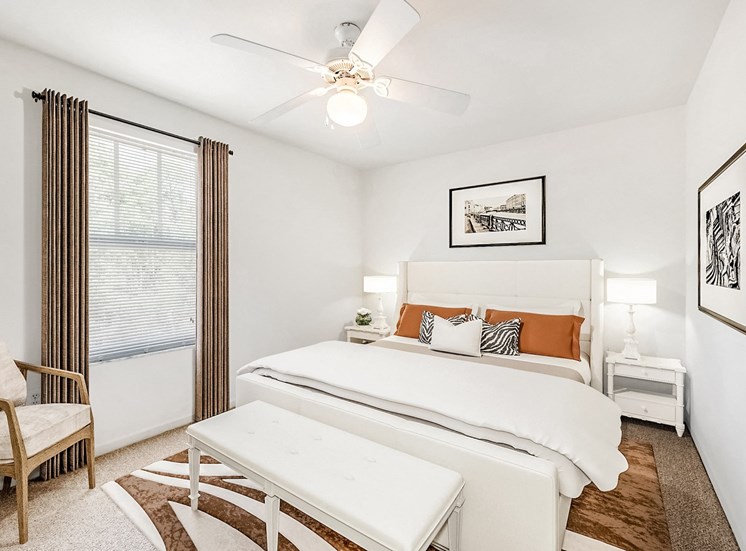 A staged bedroom with carpet, and a ceiling fan, window. Full bed with tufted headboard on top of a rug and two nightstands, and art above the bed.