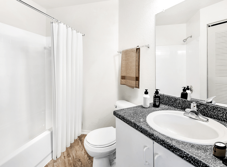 Virtually staged bathroom with large mirror, gray countertop, white cabinets and tiled tub with white shower curtain