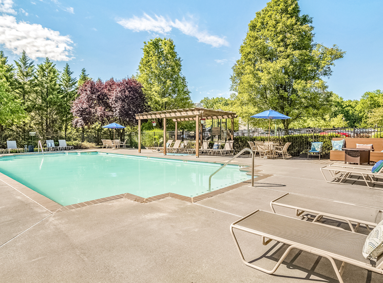 The community pool is rectangular in shape. The large sundeck area is enclosed with a black fence and features beige pool furniture including chaise lounge chairs, regular chairs, round tables, and blue umbrellas as well as a large wood-stained pergola. The exterior of the pool area is surrounded by bushes and mature trees. The community parking area is in the distance.
