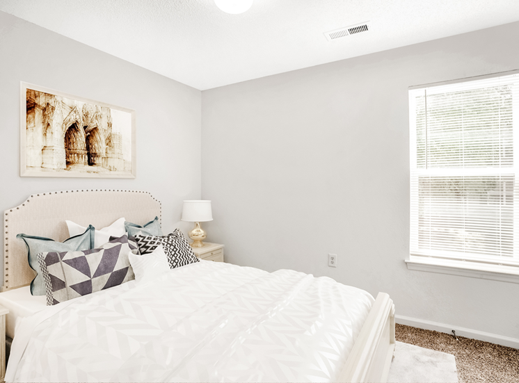 The vacant bedroom features tan carpet throughout with beige walls and a white textured ceiling. A window located on the right side of the room overlooks exterior trees. A rounded globe light sits in the center of the ceiling.