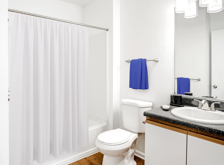 A vacant bathroom featuring white walls and a white ceiling. Brown wood-style flooring throughout. The bathroom also features a white shower/tub combo along with a black granite style bathroom vanity, white sink, and wood-colored trim surrounding the white cabinets.