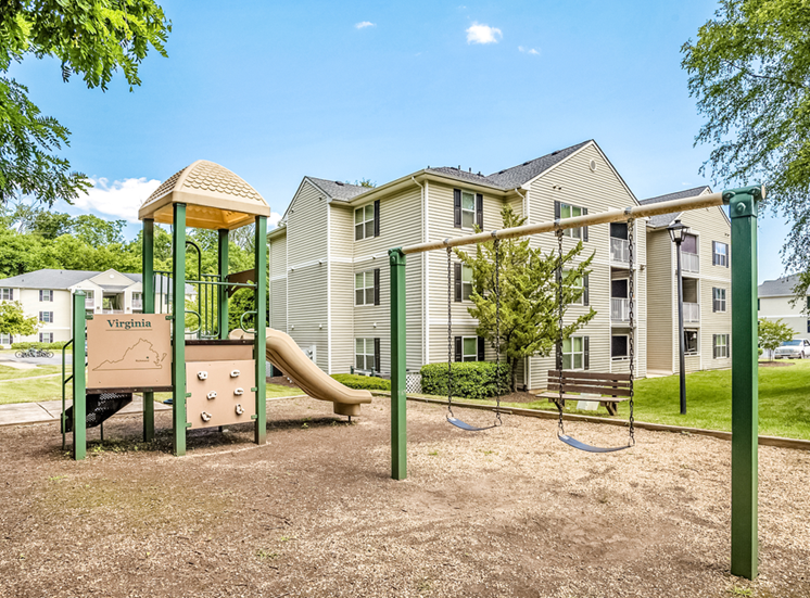The community playground features a yellow, tan, and green play structure with a slide, a climbing wall, and two seated swings. The playground is surrounded by mulch, has a bench for seating, and sits adjacent to an apartment building in the background.