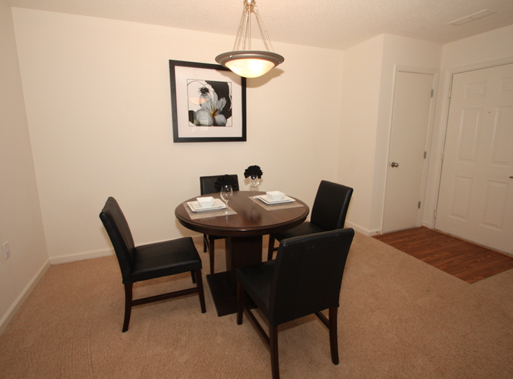 The furnished dining room has hardwood style flooring by the entry door, and carpet throughout the rest. There is a round wooden table with four black chairs positioned around it. There is a place setting for two on top of the table.