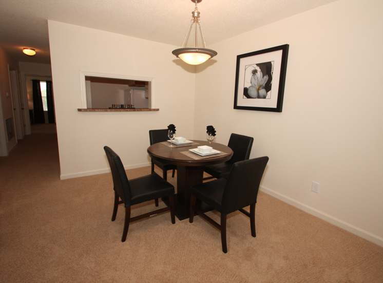 The furnished dining room has carpet throughout. There is a round wooden table with four black chairs positioned around it. There is a place setting for two on top of the table. There is a small breakfast bar that opens to the kitchen.