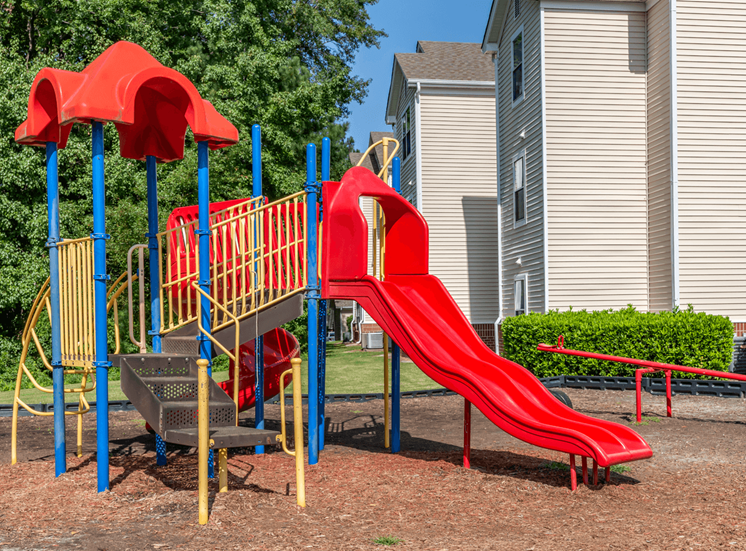 Red Yellow and Blue Playground with Red Seesaw with Building Exterior and Treeline in the Background