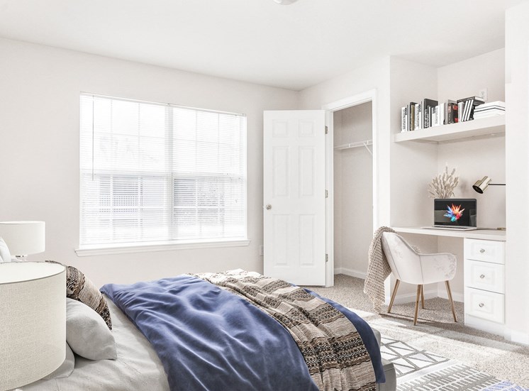 Vacant bedroom with a closet, carpet, window, and built in desk. Full bed with headboard on top of a rug in between two nightstands.  Books on shelve above desk that has laptop and lamp.