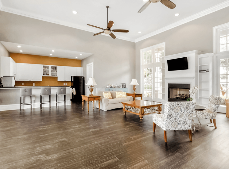 Clubhouse interior and kitchen with bar stools, wall mounted television, fireplace, wood style flooring, couch, fabric chairs, coffee table and large windows and recessed lighting