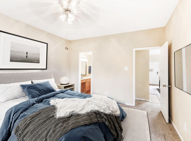 Staged bedroom with queen sized bed, art above bed, ceiling fan, nightstand, and view of bathroom and hall.