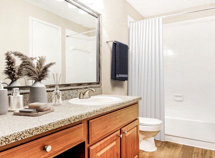 Staged brown speckled counter top with décor on top of it and brown cabinets with wood style flooring. Framed mirror above sink with view of bathtub in the background.