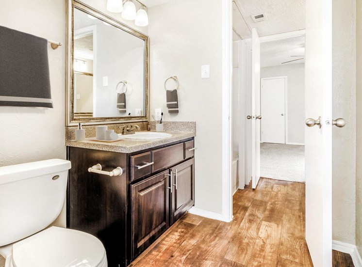 Staged brown speckled counter top with décor on top of it and brown cabinets with wood style flooring. Framed mirror above sink with view od bathtub in the background.