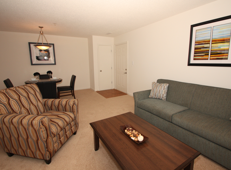 The furnished model home has hardstyle flooring in the entry by the front door with carpet in the living room and dining room. This floorplan is an open. There is a gray upholstered couch, a tan, blue, and red patterned chair, a dark wooden coffee table, a round dining table with 4 chairs and framed artwork hanging on the walls.
