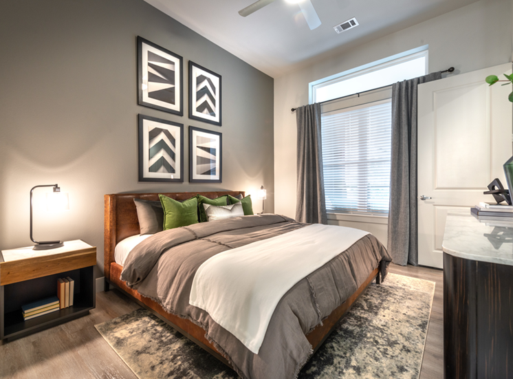 Staged bedroom with bed, accent rug, wood style flooring nightstands with lamps, gray accent wall with art, ceiling fan and large window with blinds and curtain