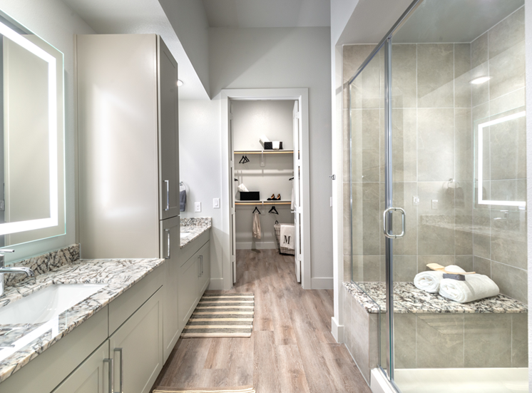 Staged bathroom with dual vanities, granite countertops, taupe cabinetry, walk in shower with bench, wood style flooring and direct access to walk in closet
