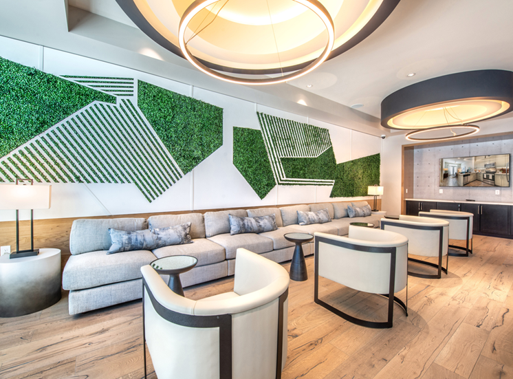Clubhouse lounge with long couch and chairs with unique green wall art and large circular light fixtures with wall mounted television